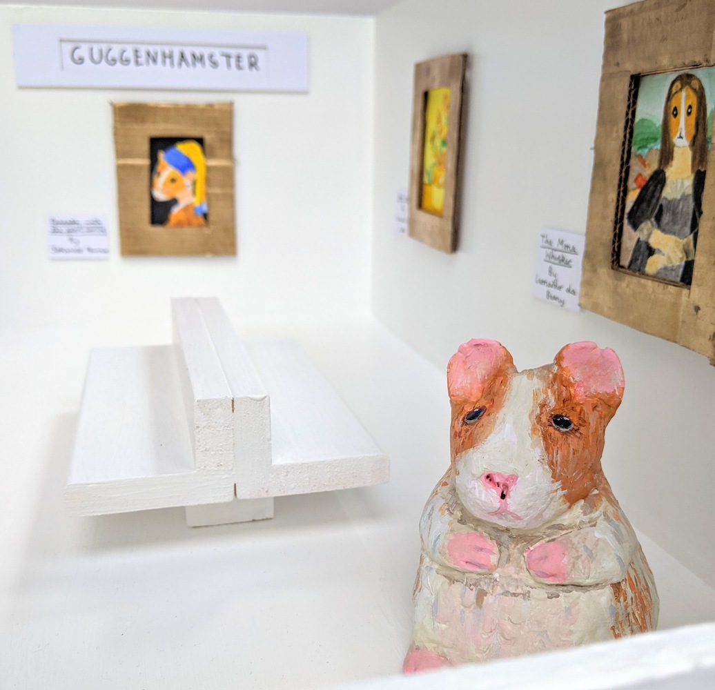 This artwork is a white box with an open top, featuring 6 small famed paintings. The paintings include homages to famous artworks, that have been reimagined with a hamsters face. The box is designed to look like a modern art gallery, with white walls and floor, and a white bench in the centre. There is a ceramic hamster, painted white with an orangey brown head in the gallery gazing at one of the artworks. There are plaques with the new names of the paintings underneath. These include The Squeak (aka The Scream), Self Pawtrait (A Frida Kahlo homage), Pop Pancake (in the style of Andy Warhol), Pancake with the Pearl Earring (in the style of Girl with the Pearl Earring), Small Sunflowers and The Mona Whister (in the style of The Mona Lisa).