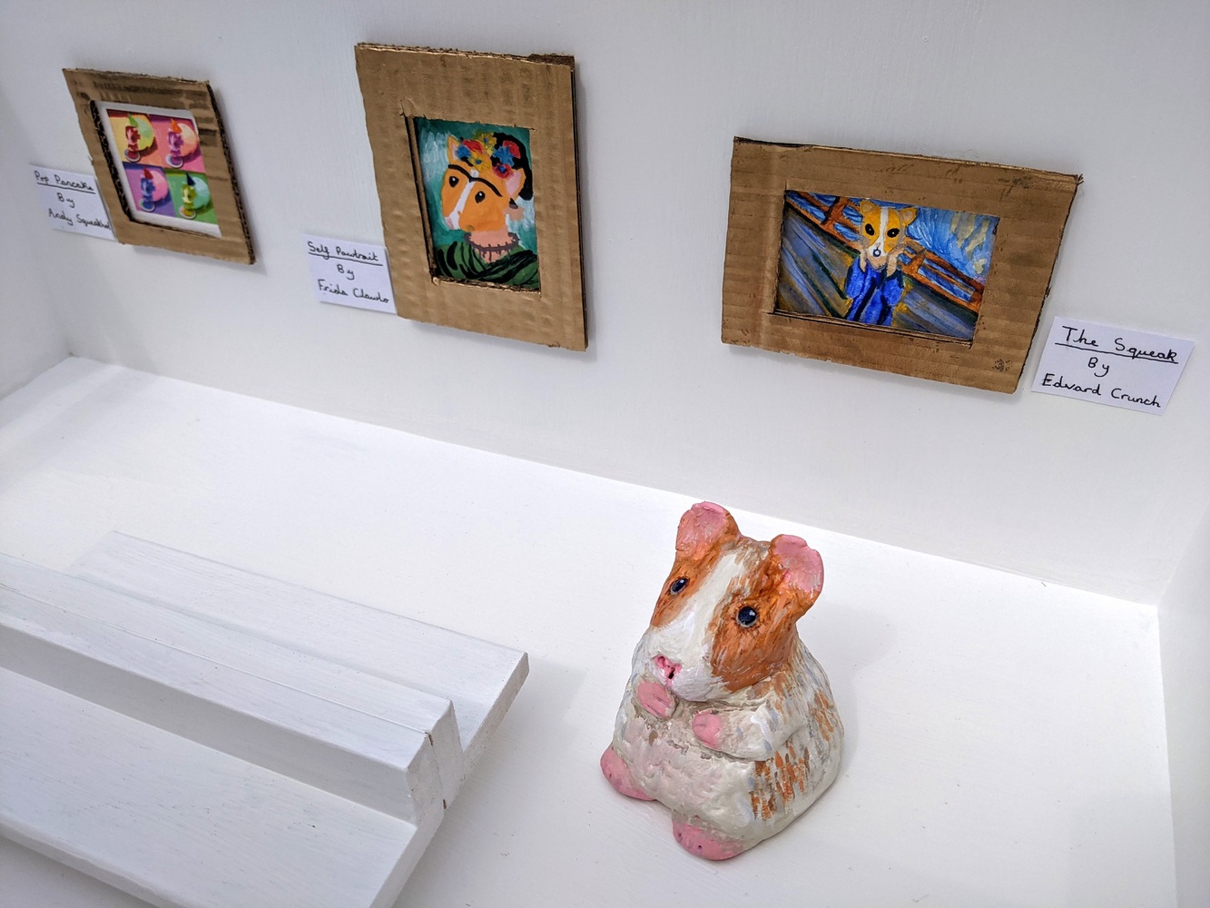 This artwork is a white box with an open top, featuring 6 small famed paintings. The paintings include homages to famous artworks, that have been reimagined with a hamsters face. The box is designed to look like a modern art gallery, with white walls and floor, and a white bench in the centre. There is a ceramic hamster, painted white with an orangey brown head in the gallery gazing at one of the artworks. There are plaques with the new names of the paintings underneath. These include The Squeak (aka The Scream), Self Pawtrait (A Frida Kahlo homage), Pop Pancake (in the style of Andy Warhol), Pancake with the Pearl Earring (in the style of Girl with the Pearl Earring), Small Sunflowers and The Mona Whister (in the style of The Mona Lisa).