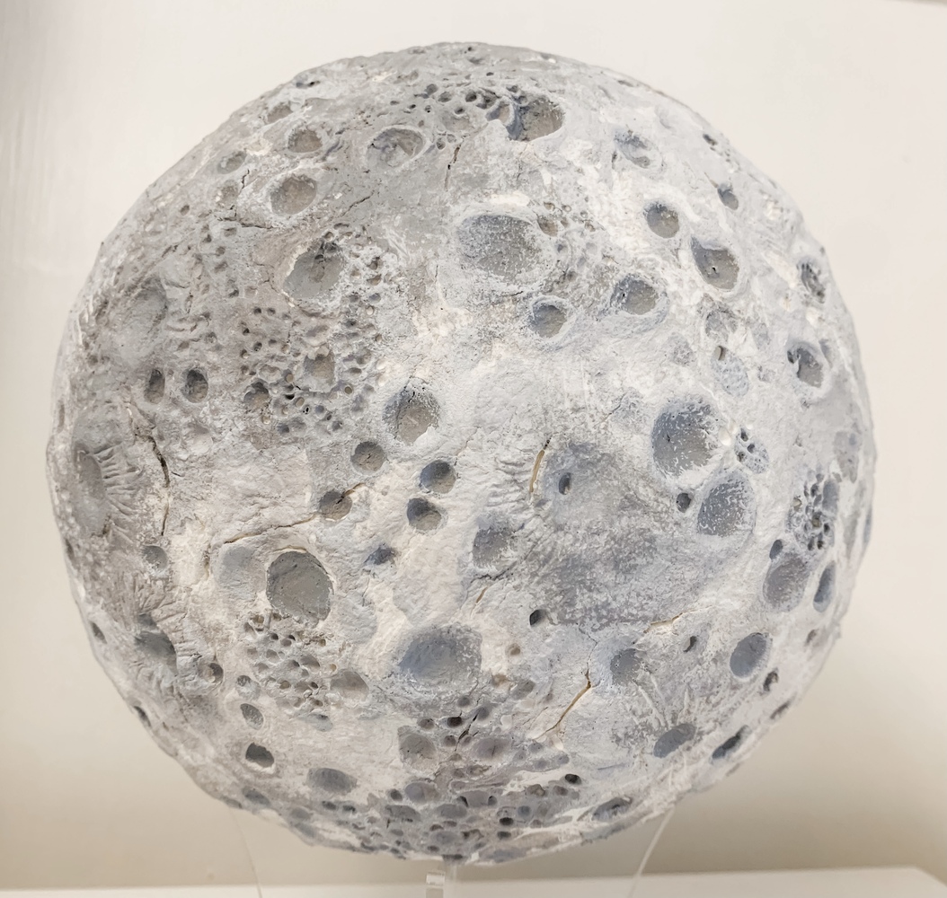 Shades of grey and white reveal the textured surface of the lunar surface. Craters of varying sizes, depths and origins are embedded in the ancient landscape. An ancient lava plain, Lacus Mortis or "lake of death", appears in the northeastern region of the face of the moon.